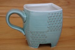 Pseudocubic mug with raised feet and hexagonal lattice motif.  Thrown then altered porcelain with celadon glaze and hematite slip, cone 6 oxidation.