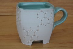 Pseudocubic mug with raised feet and hexagonal lattice motif.  Thrown then altered porcelain with celadon glaze and hematite slip, cone 6 oxidation.